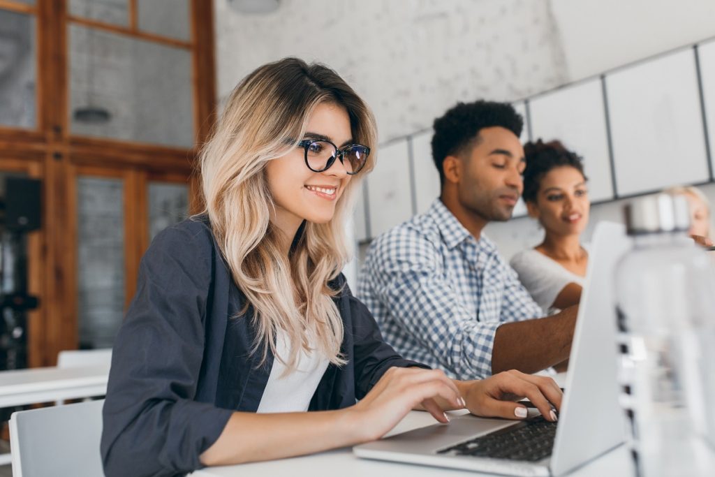 Beautiful Curly Female Freelancer With Cute Manicure Using Laptop And Smiling Indoor Portrait Of Blonde Secretary Sitting Beside African Coworker In Blue Shirt